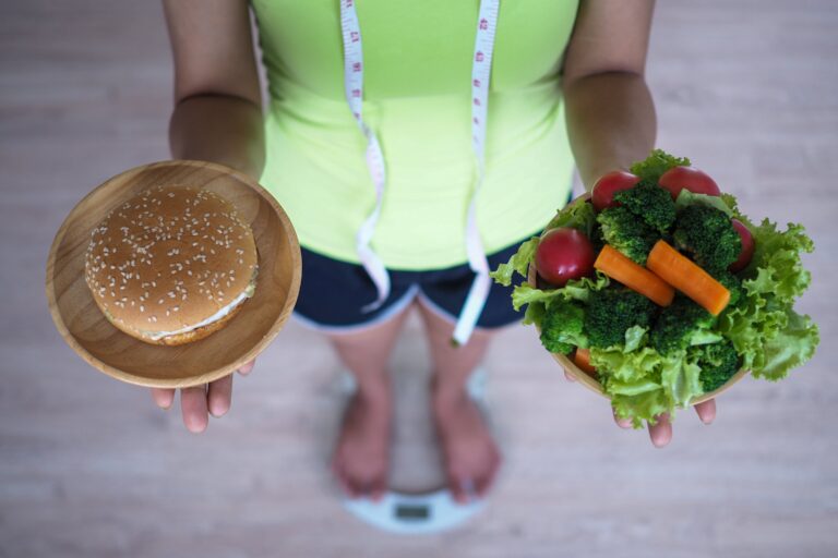 Eine Person steht auf einer Waage, hält in der einen Hand einen Teller mit einem Burger und in der anderen eine Schüssel mit Gemüse und überlegt, welches Essen sie auswählen soll.