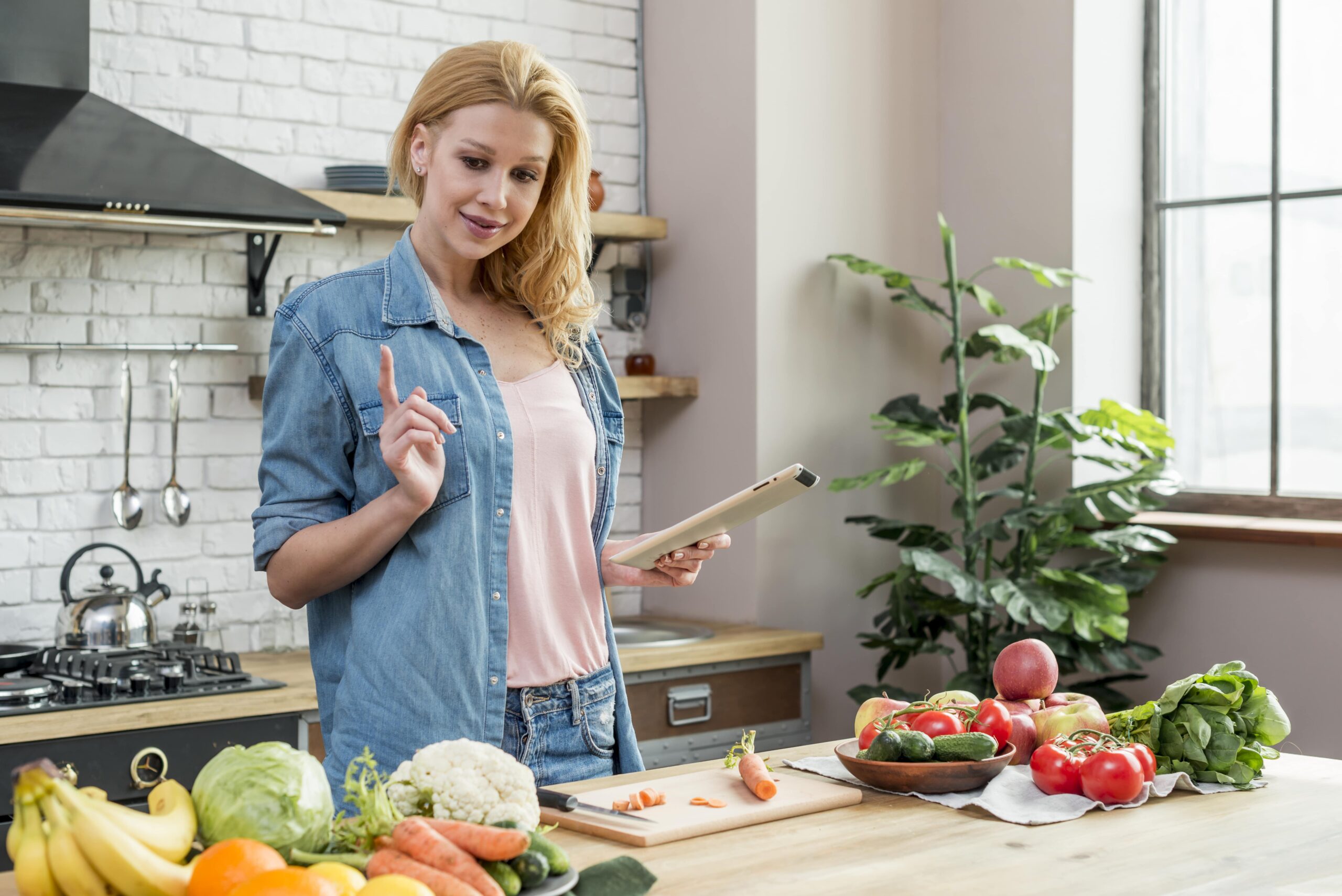 Eine Frau in einer Küche hält in einer Hand ein Tablet und macht mit der anderen eine Zeigegeste, umgeben von einer Auswahl an frischem Obst und Gemüse.