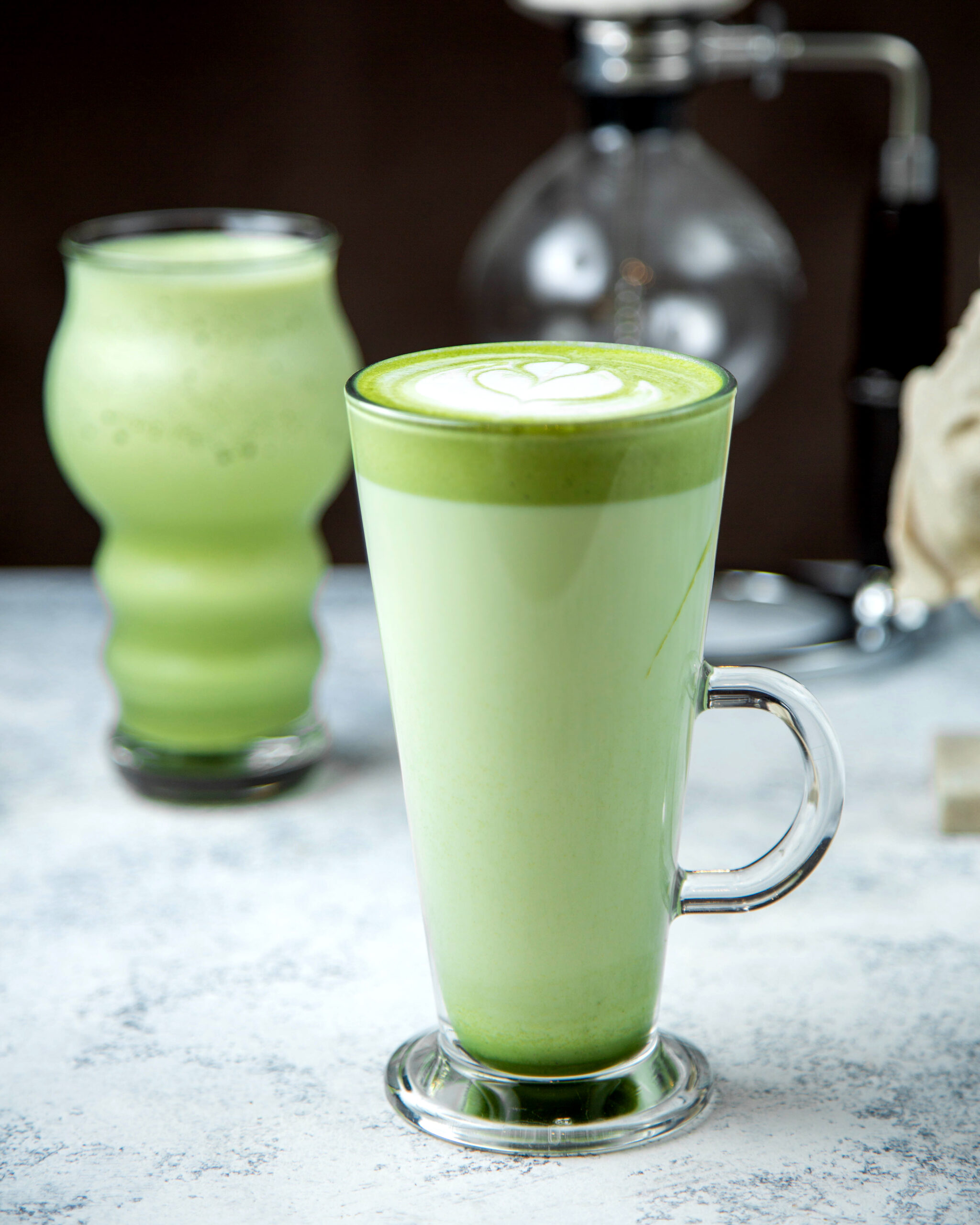 Ein Glas selbst gemachter grüner Shake, im Hintergrund ist ein weitere Shake und beide stehen auf einer grauen Oberfläche.