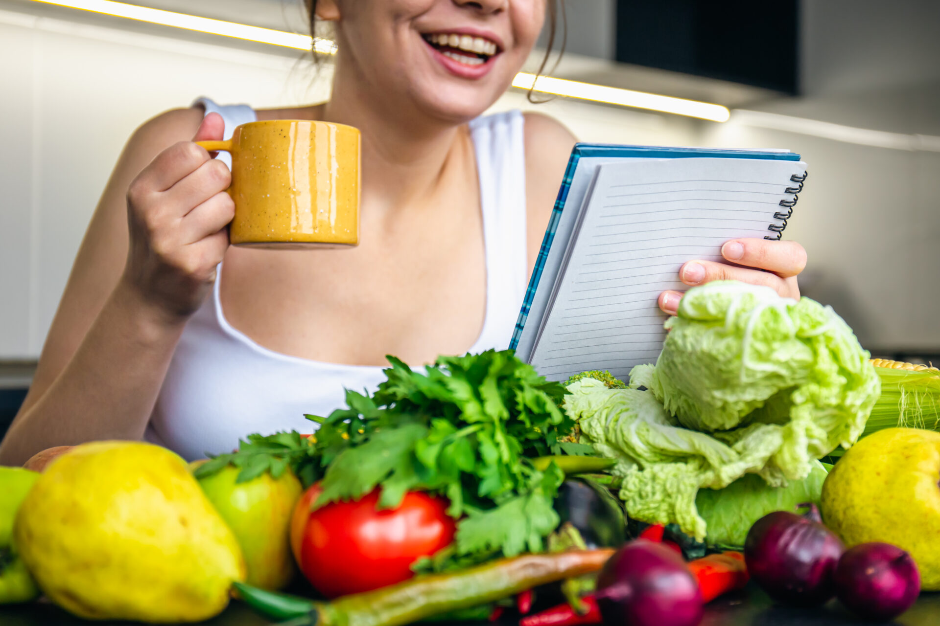 Frau liest in einer Küche in einem Notizbuch und trinkt dabei aus einer Tasse, vor ihr liegt verschiedenes Gemüse