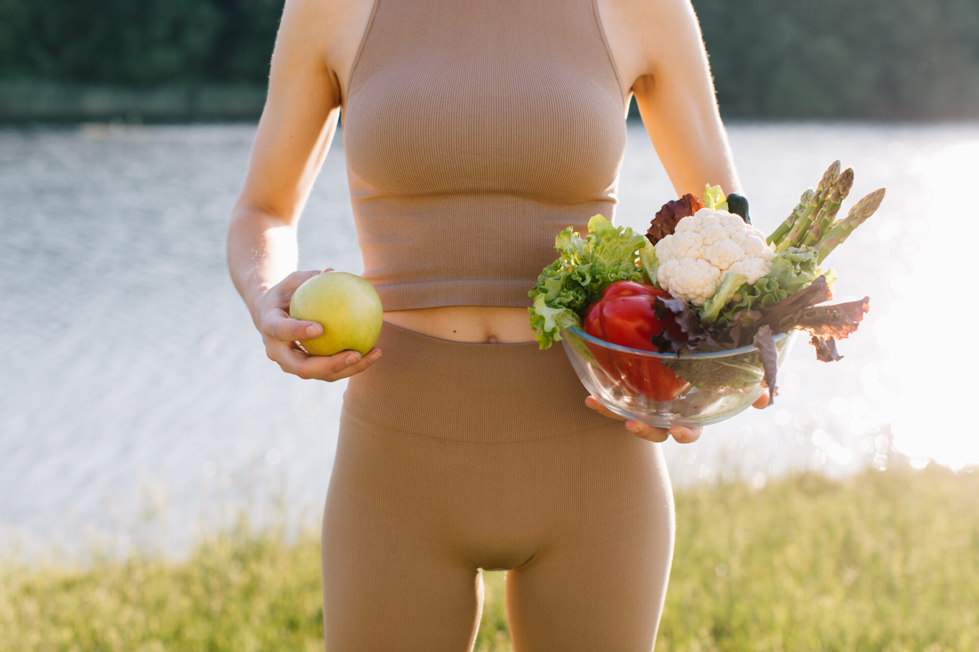 Frau hält in der einen Hand einen Apfel und in der anderen frisches Gemüse nahe dem Bauch, an einem sonnigen Tag vor einem See