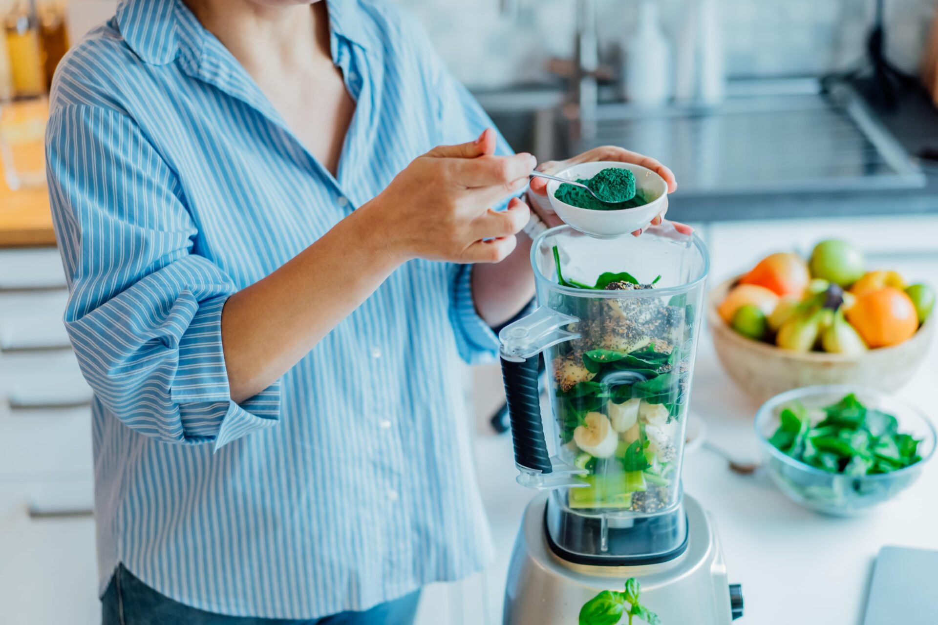 Eine Frau im gestreiften Hemd gibt in einer Küche einen Löffel grünes Spirulinapulver in einen mit Obst und Gemüse gefüllten Mixer.