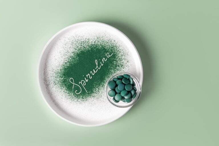 Ein weißer Teller mit grünem Spirulinapulver, in dem das Wort „Spirulina“ steht und daneben steht eine kleine Glasschale gefüllt mit grünen Spirulinatabletten vor einem grünen Hintergrund.
