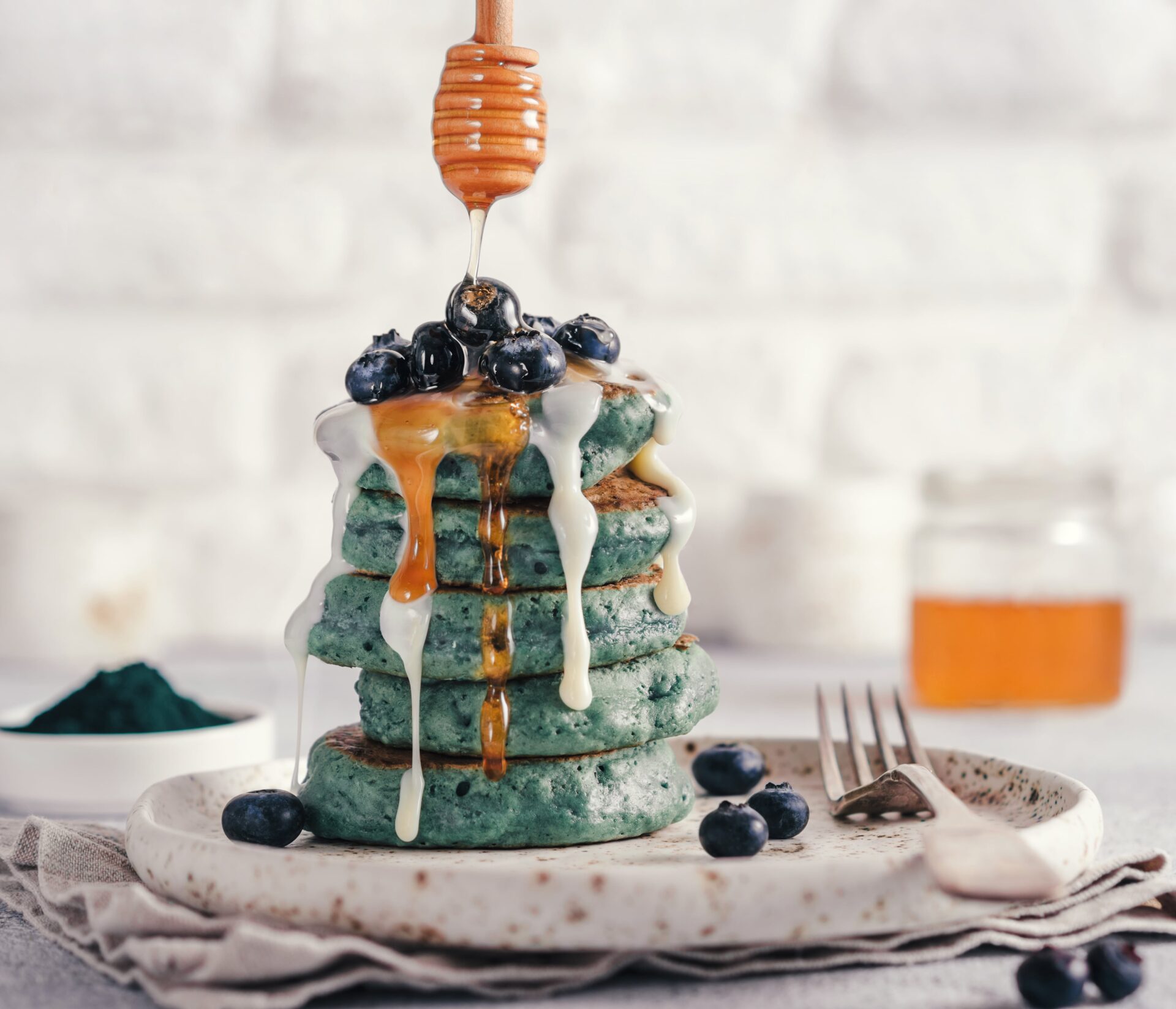Ein Stapel blauer Pfannkuchen mit Blaubeeren, Honig und weißer Soße liegt auf einem Teller, daneben liegt eine Gabel. Von oben wird Honig darübergeträufelt. Im Hintergrund ist ein Glas Honig verschwommen.