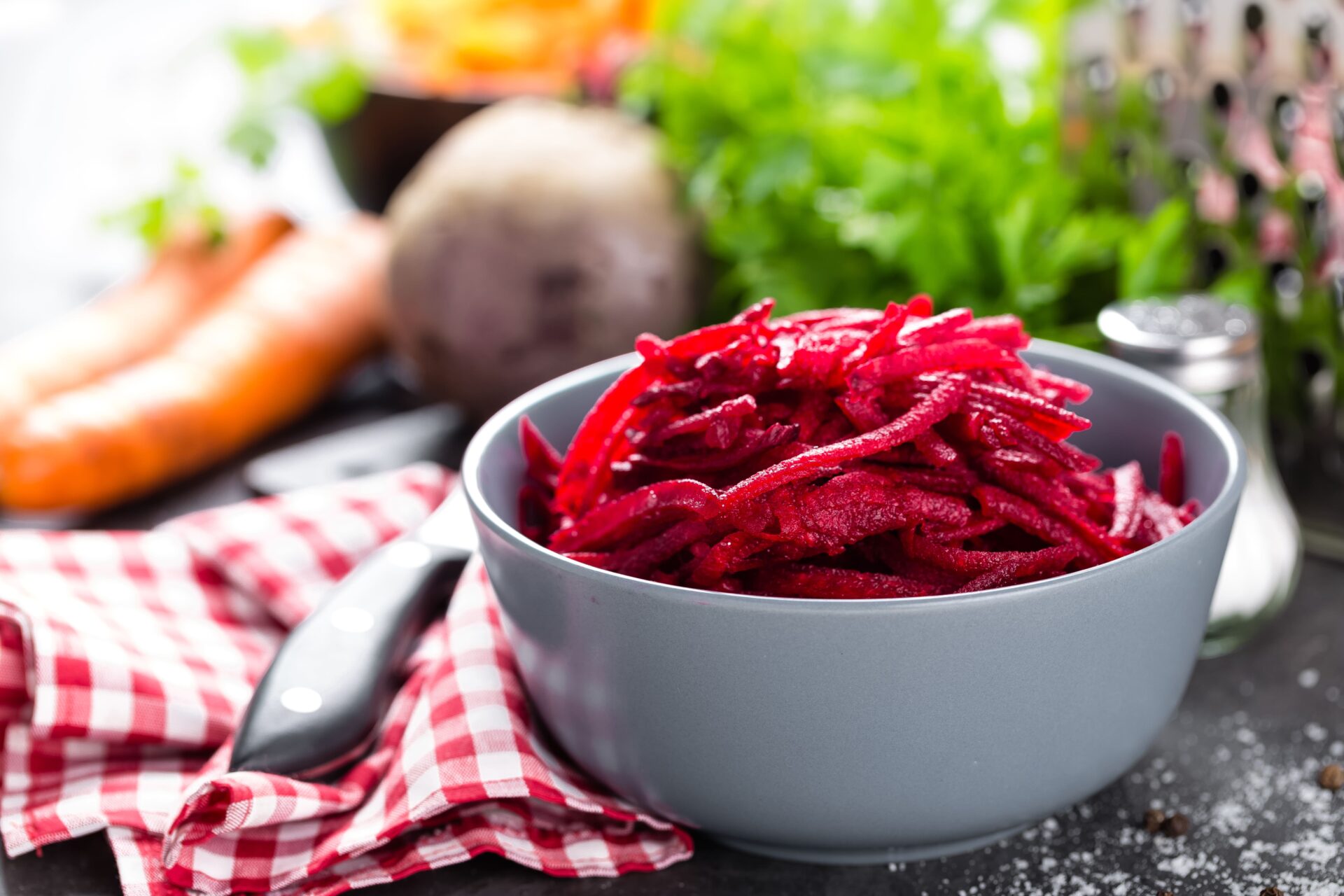 Auf einem Tisch steht eine graue Schüssel gefüllt mit leckerem Rote-Bete-Salat, im Hintergrund ein kariertes Tuch, ein Messer und verschiedene Gemüsesorten.