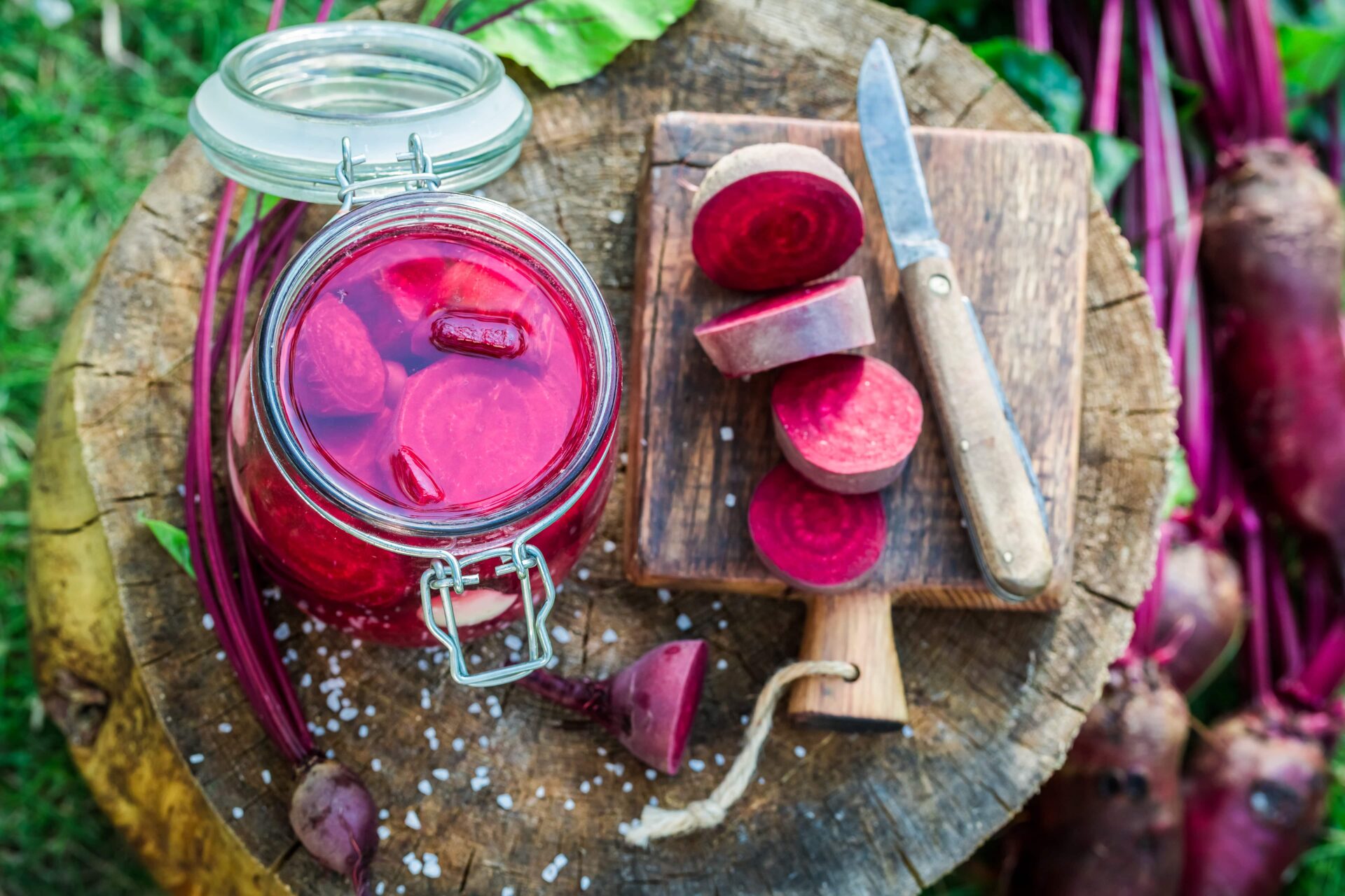 Ein Glas mit eingelegter Roter Bete auf einer Holzoberfläche neben geschnittener Roter Bete auf einem Schneidebrett, einem Messer und frischen roten Rüben mit Stielen und Blättern.