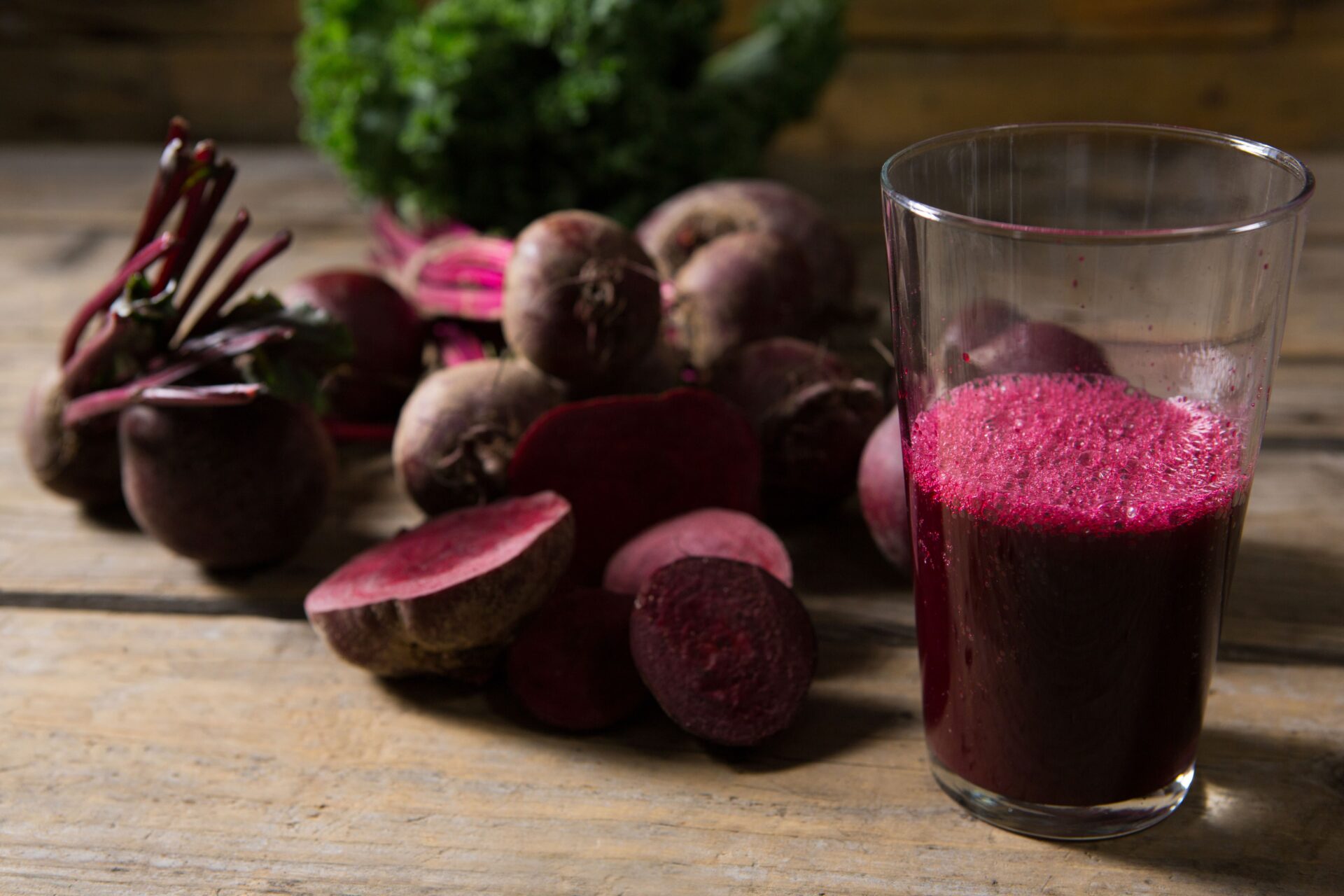 Ein Glas Rote-Bete-Saft steht auf einem Holztisch neben ganzen und geschnittenen roten Rüben, im Hintergrund ist Blattgemüse zu sehen.