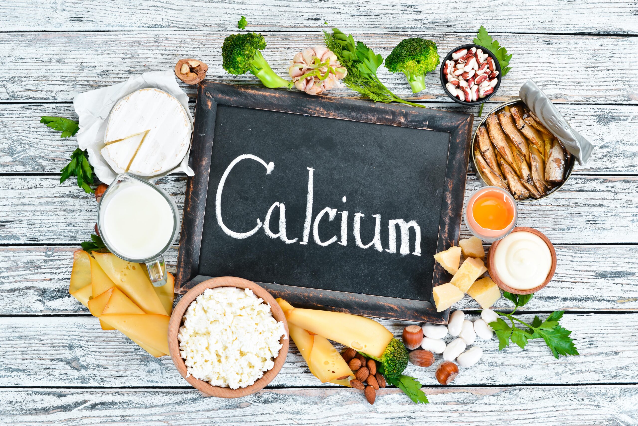 Eine Tafel mit der Aufschrift „Calcium“ ist umgeben von verschiedenen kalziumreichen Lebensmitteln wie Milch, Käse, Brokkoli, Fisch, Mandeln und Joghurt auf einem Holztisch.