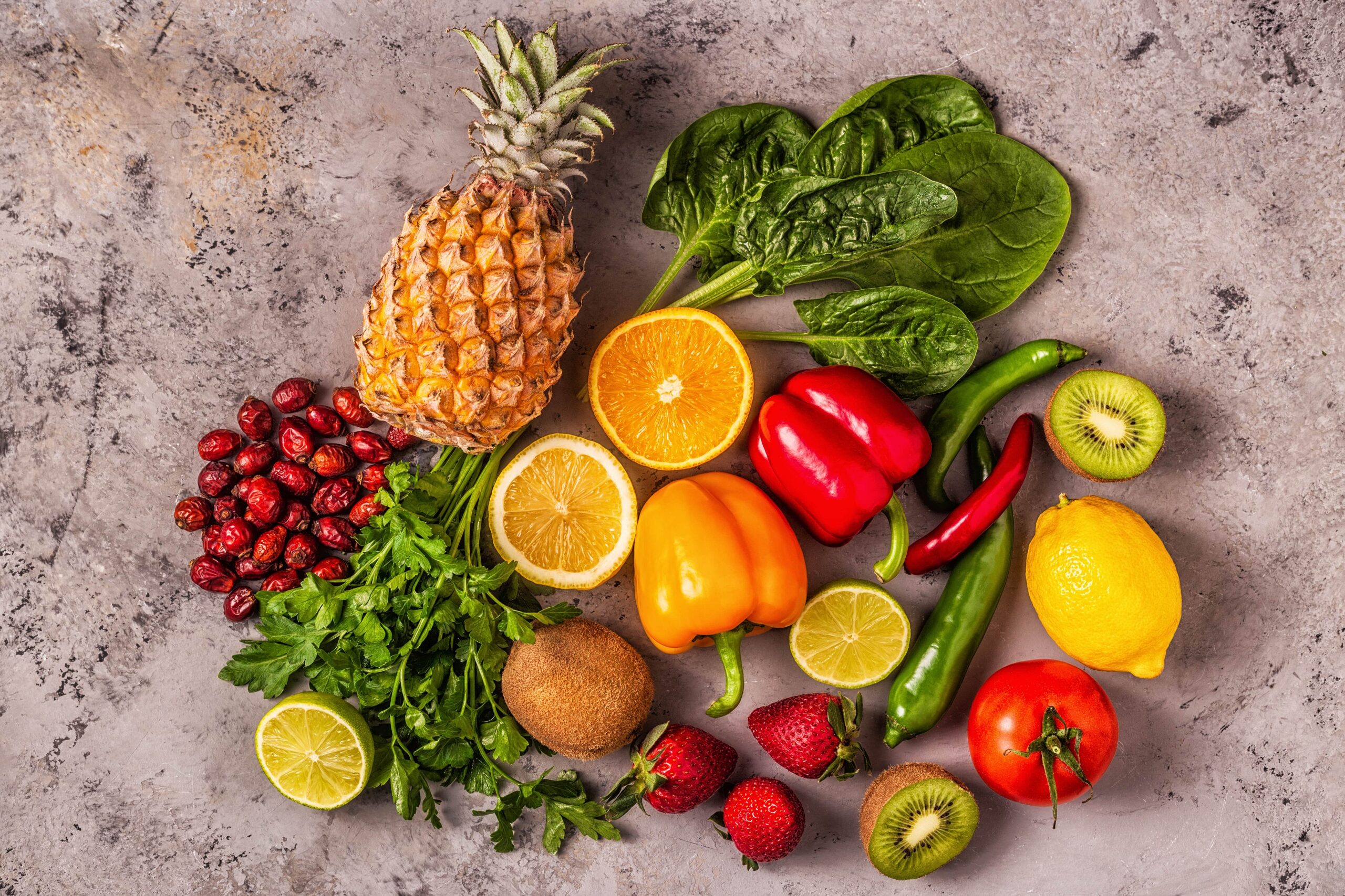 Verschiedene Obst- und Gemüsesorten auf einer strukturierten Oberfläche angeordnet, darunter Ananas, Paprika, Spinat, Tomaten, Kiwi, Zitrone, Orange, Erdbeeren, Kirschen, Limette und Chilischoten.