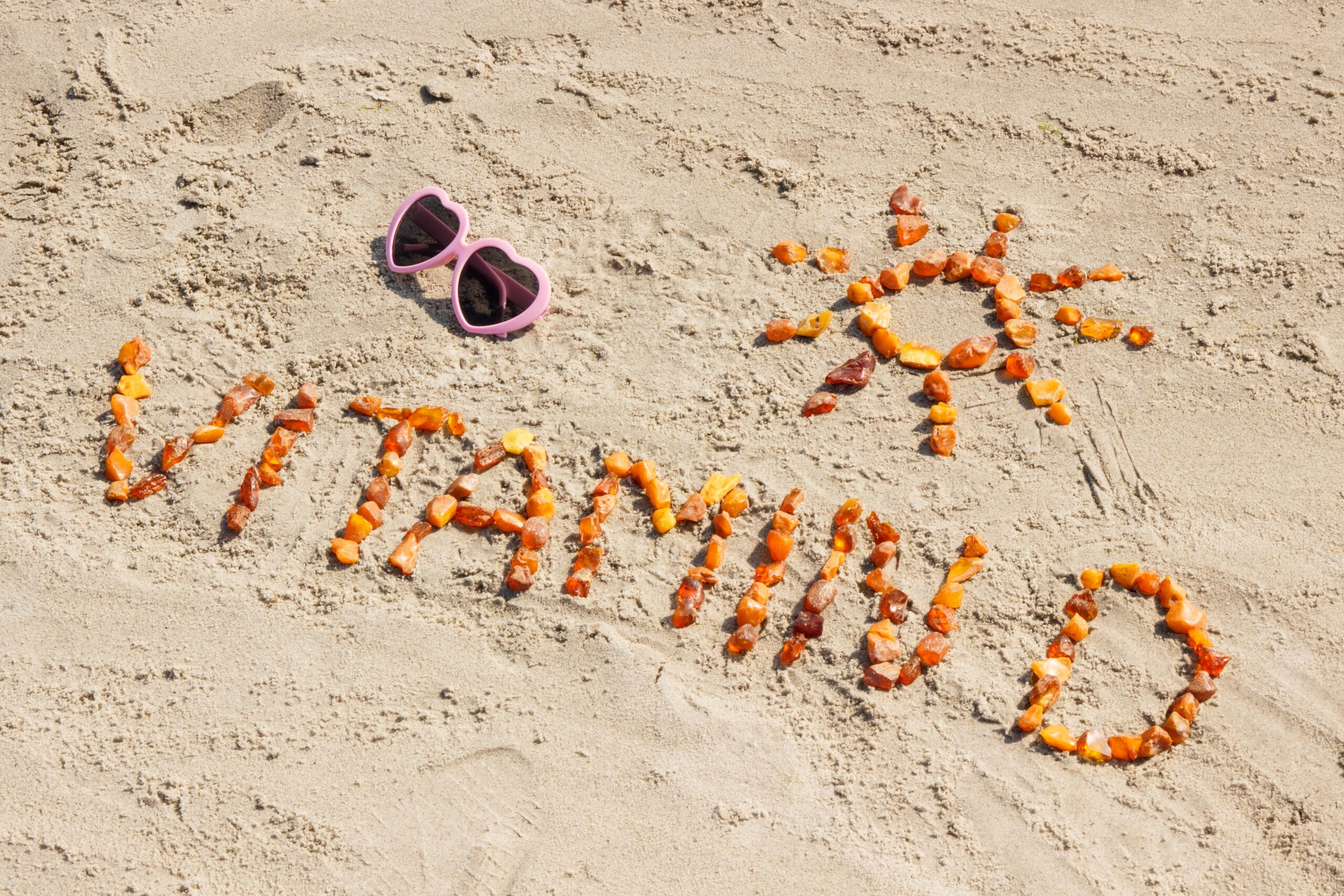 „Vitamin D“ ist aus kleinen Steinen auf einem Sandstrand geformt, mit einer Sonne, ebenfalls aus Steinen, und einer rosa Sonnenbrille über dem Text.