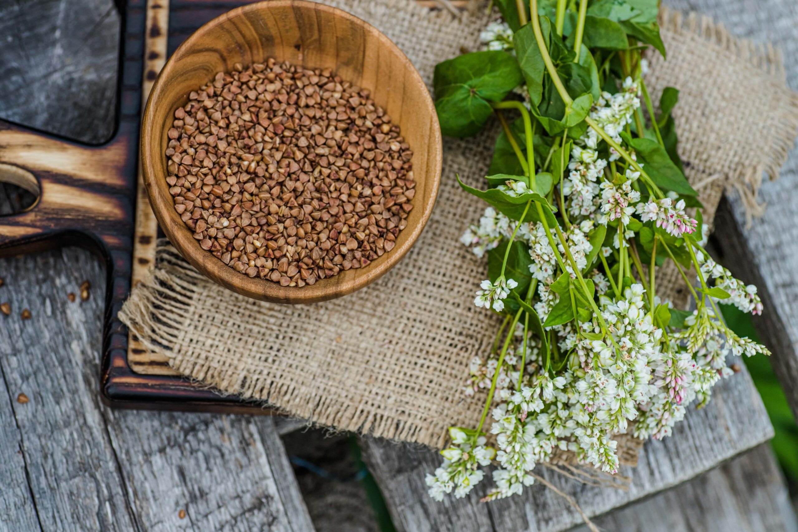 
Eine Holzschale mit Buchweizengrütze steht auf Sackleinen neben einem Strauß weißer Blumen auf einer rustikalen Holzoberfläche.