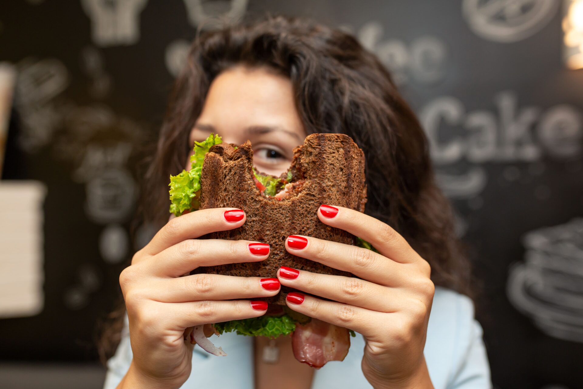 Person mit rotem Nagellack hält ein Sandwich mit dunklem Brot und Salat und bedeckt ihr Gesicht vor einer Kreidetafel mit der Speisekarte.