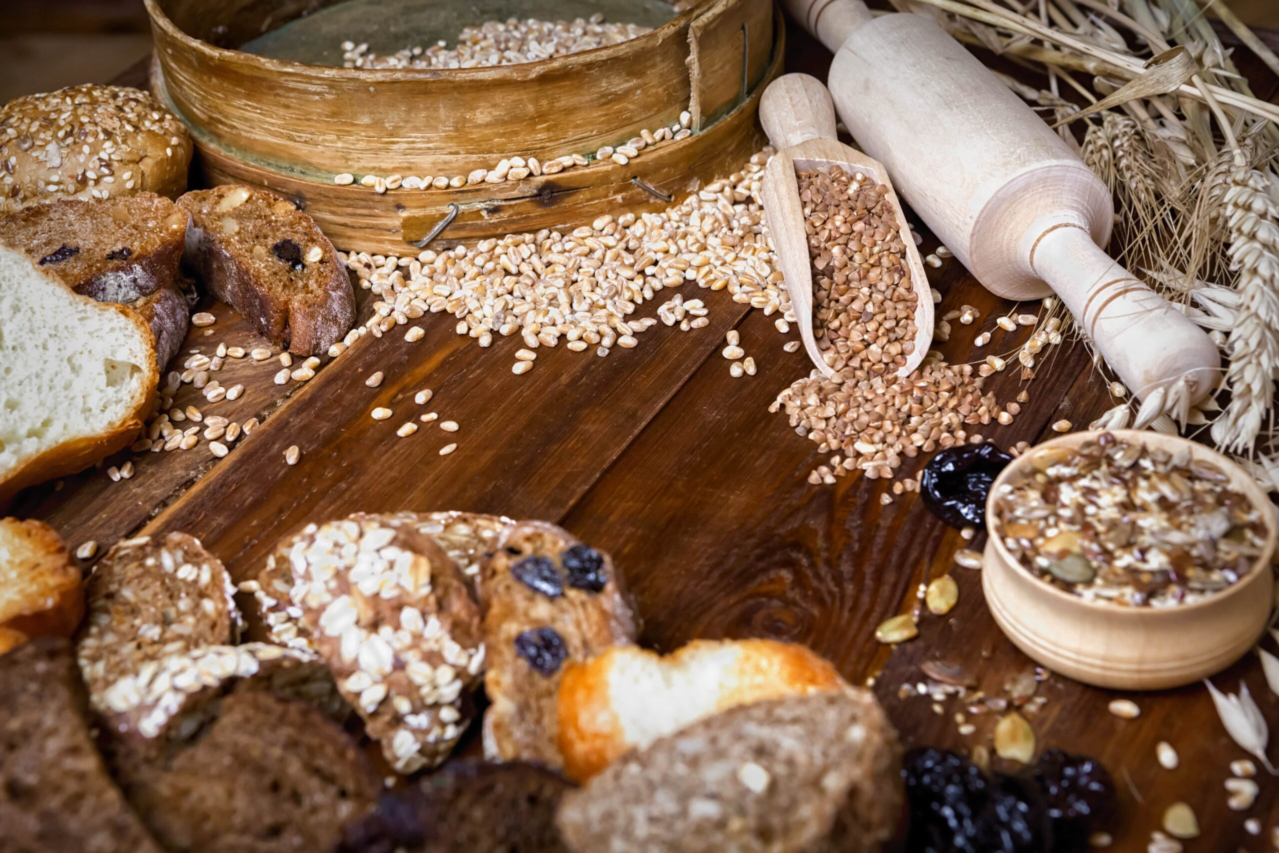 Verschiedene Brotsorten, Körner und Samen werden zusammen mit einem Nudelholz und Weizenhalmen auf einem Holztisch präsentiert.