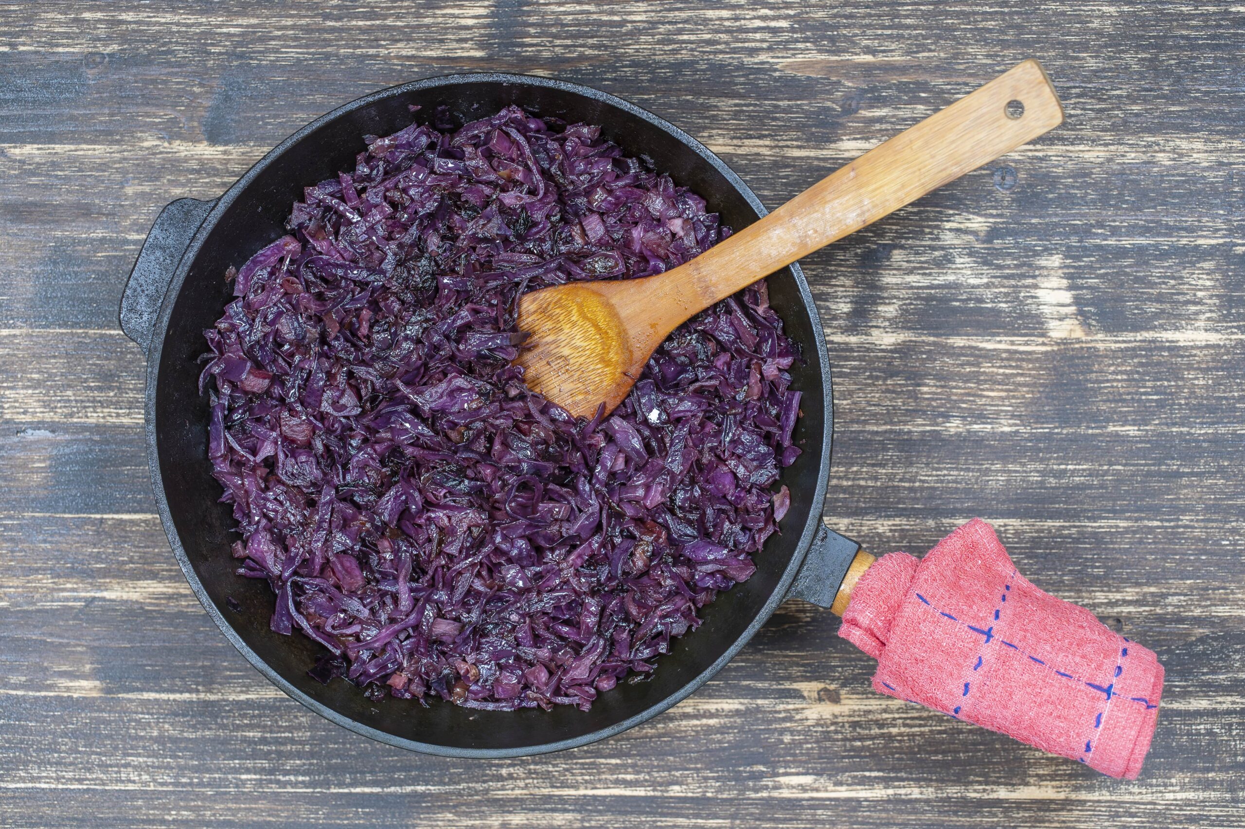 Gekochter Rotkohl in einer schwarzen Pfanne mit einem Holzlöffel, auf einer Holzoberfläche platziert.