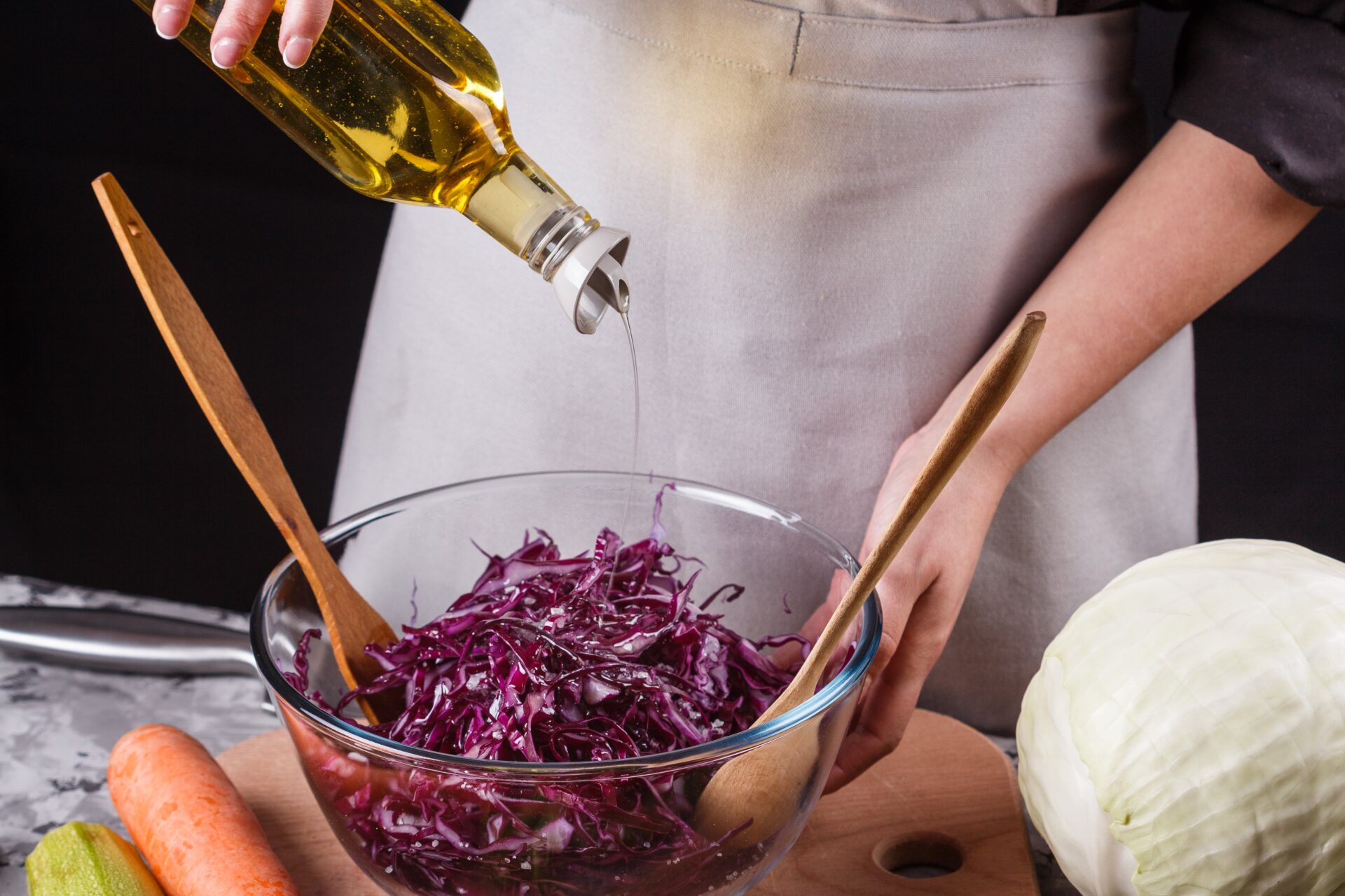 Eine Person gießt mit Holzbesteck Olivenöl in eine Schüssel mit zerkleinertem Rotkohl, neben ihr liegen eine Karotte und ein ganzer Kohl auf einem Schneidebrett.