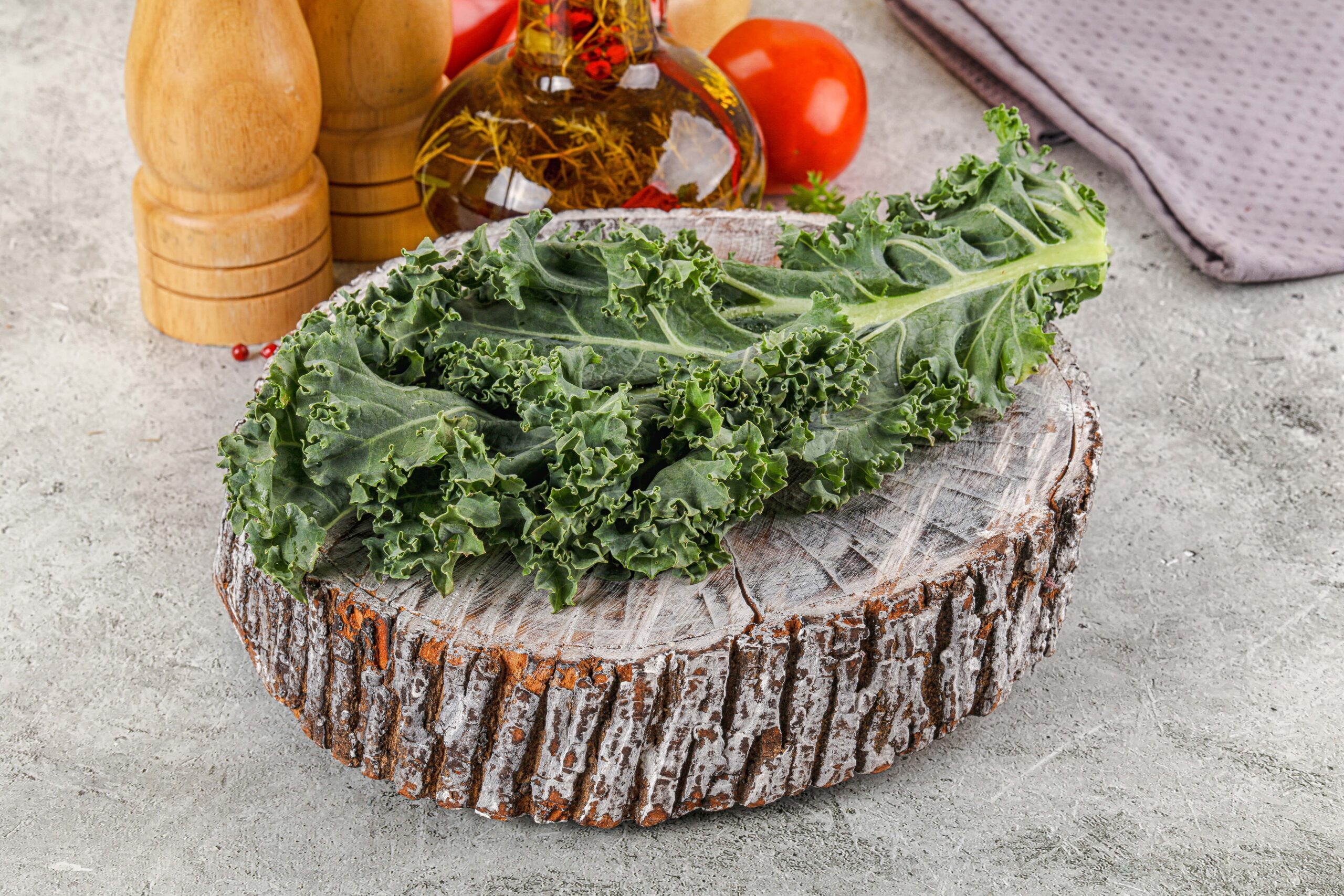 Ein Grünkohlblatt auf einem runden Holzbrett mit Pfeffermühlen, Ölflasche, Tomate und einem grauen Tuch im Hintergrund.