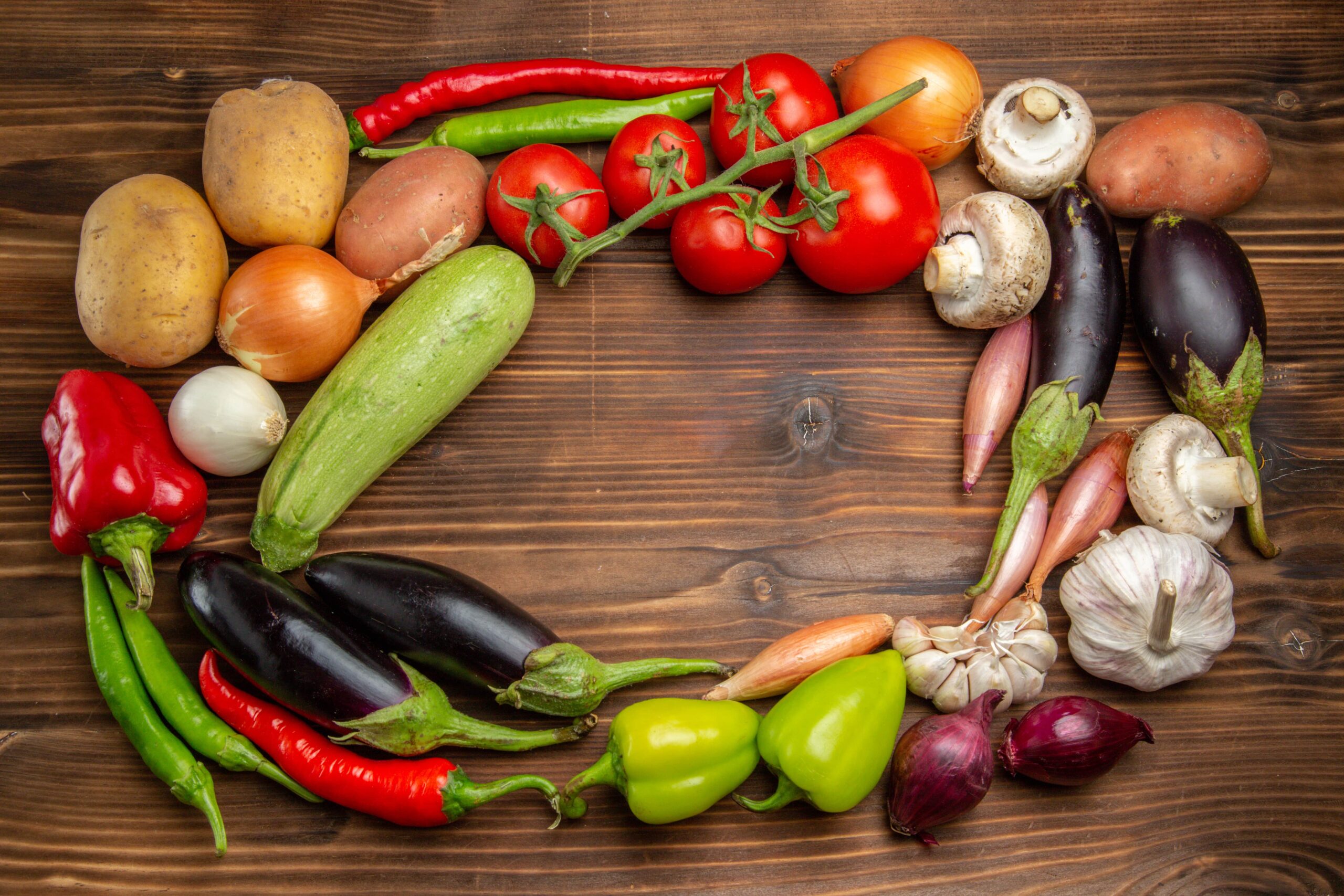 
Eine Auswahl an Gemüse, darunter Tomaten, Paprika, Kartoffeln, Zwiebeln, Auberginen und Knoblauch, kreisförmig auf einer Holzoberfläche angeordnet.