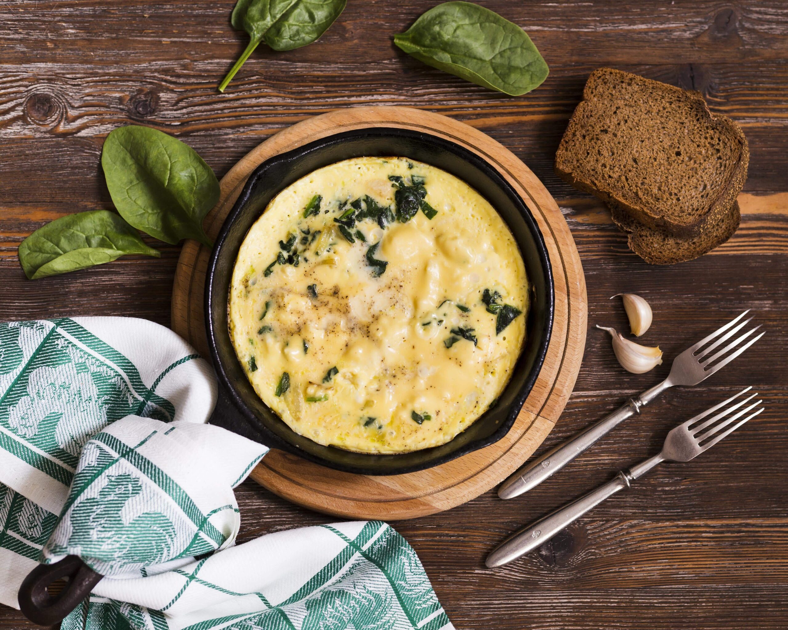 Pfanne mit einer Käse-Lauch-Suppe auf einem Holzbrett, dazu Spinatblätter, zwei Scheiben Brot, Knoblauchzehen, zwei Gabeln und ein gemustertes Handtuch auf einer Holzoberfläche.