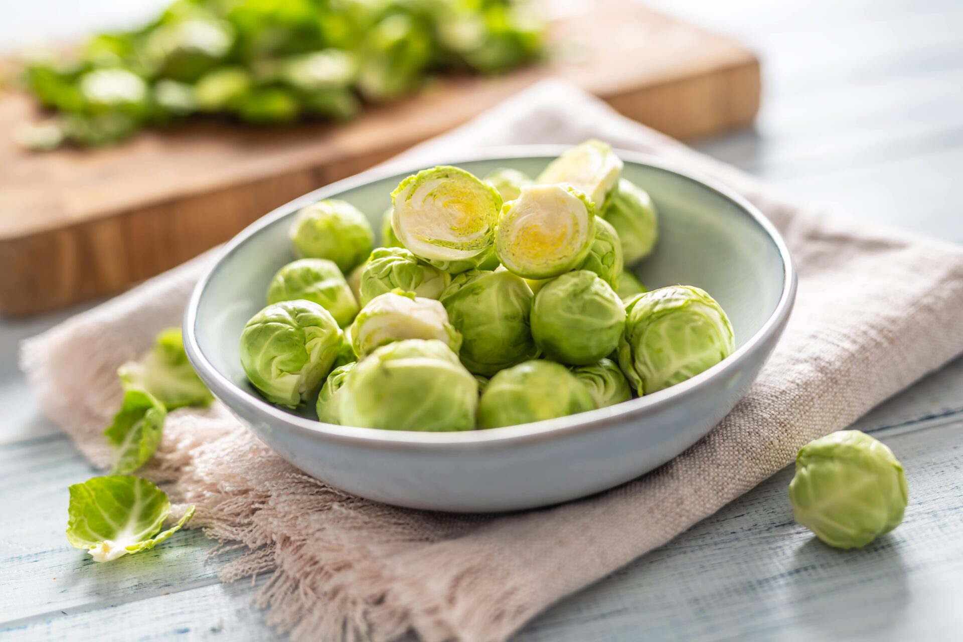 Eine Schüssel mit frischem Rosenkohl auf einer Stoffserviette, im Hintergrund ein Holzschneidebrett und die abgezupften Blätter vom Rosenkohl.